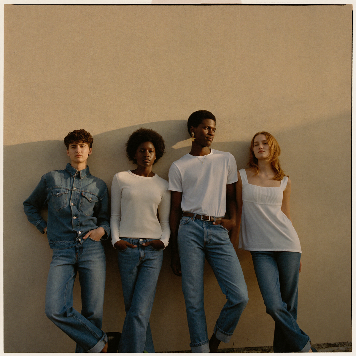 Four people leaning against a sunlit wall, all wearing blue denim